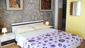 Un dormitorio con pared de piedra, una cama con cubrecama floral púrpura, dos cuadros, una mesita de noche con una lámpara y un espejo en la pared.