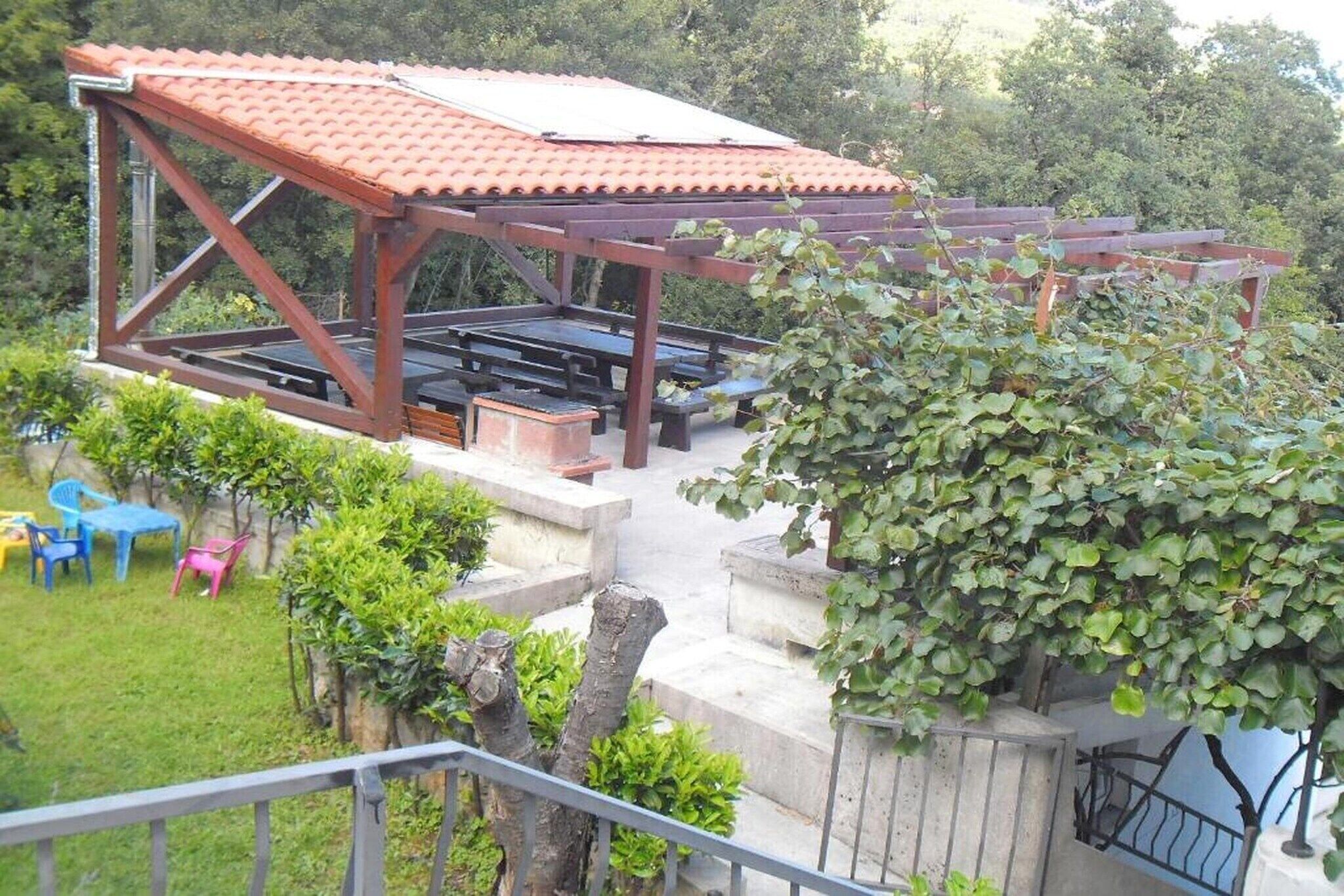 A covered outdoor seating area with benches and tables, surrounded by greenery and a small garden.
