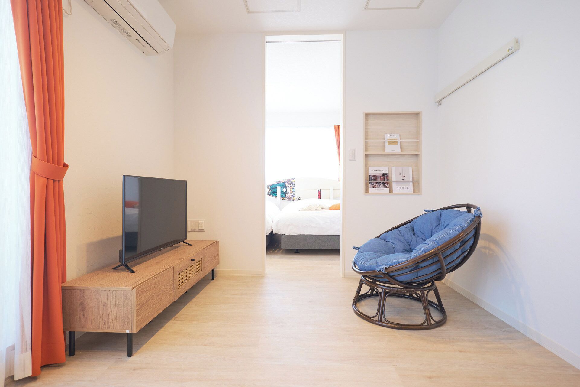 A room with a TV on a wooden cabinet, a wicker chair, a bed with a white headboard, and a shelf with framed pictures.
