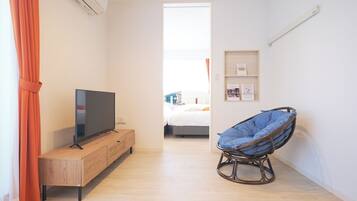 A room with a TV on a wooden cabinet, a wicker chair, a bed with a white headboard, and a shelf with framed pictures.