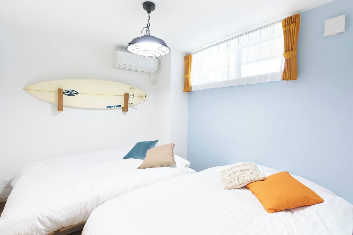 A bedroom with two beds, a surfboard on the wall, and a window with curtains.