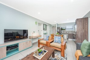 Ein modernes Wohnzimmer mit Flachbildfernseher, Holzmöbeln, einem Couchtisch und einer Küche im Hintergrund.