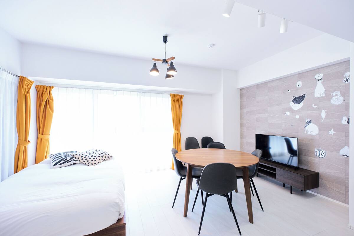 A modern hotel room with a bed, a dining table with chairs, a TV, and a wall with decorative stickers.