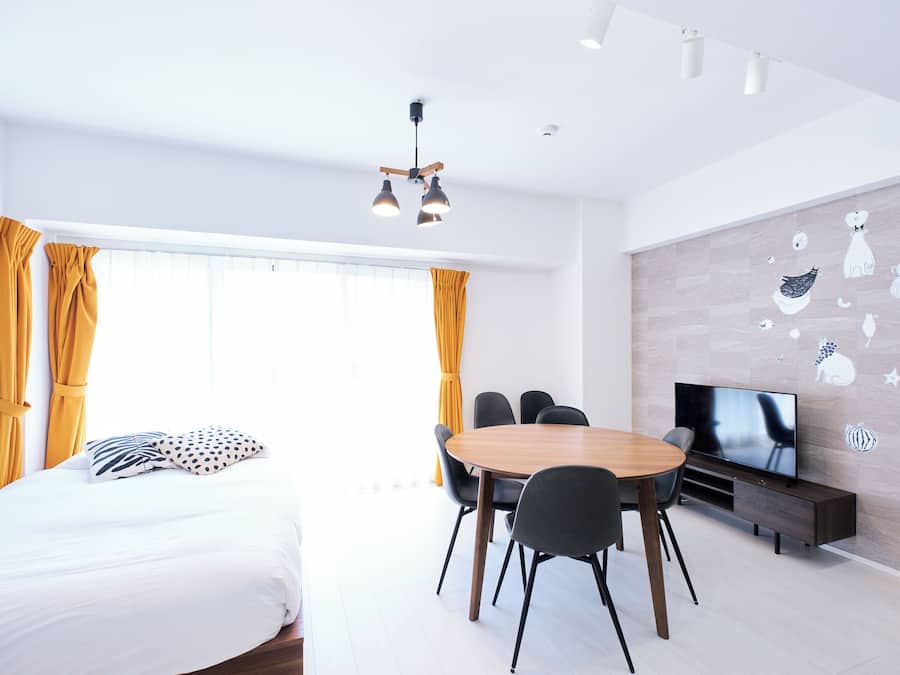 A modern hotel room with a bed, a dining table with chairs, a TV, and a wall with decorative stickers.