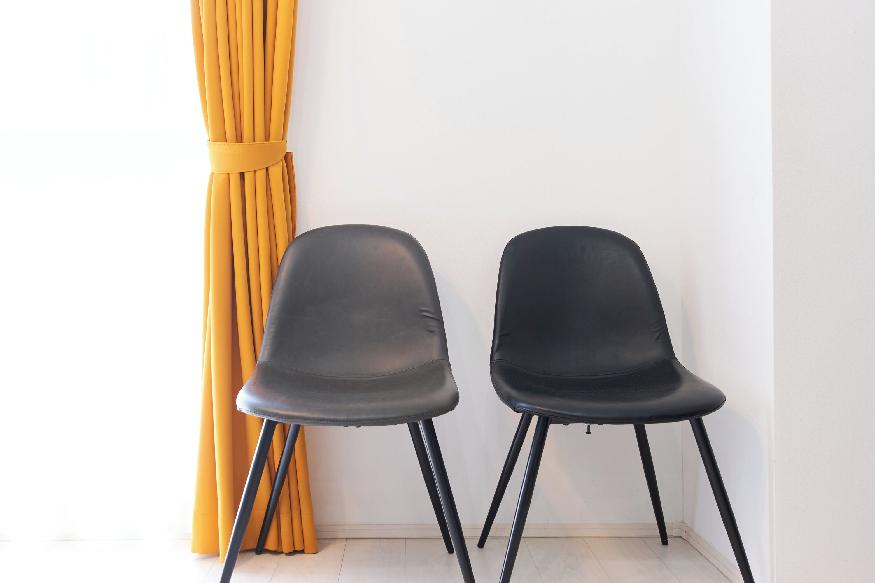 Deux chaises noires à pieds noirs, placées contre un mur blanc, avec un rideau jaune.