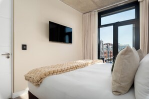 A hotel room with a bed, a television mounted on the wall, and a window with a view of buildings. - The Signature | Designer De Waterkant Retreat (Cape Town)