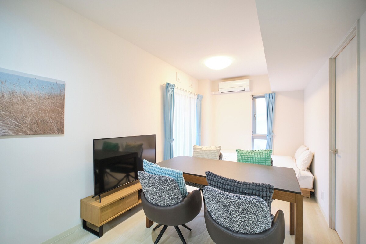 Ein modernes Wohnzimmer mit Flachbildfernseher, einem Esstisch, zwei Stühlen, einer Couch und einem Bild an der Wand.