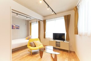 A modern room with a bed, a yellow chair, a glass coffee table, a TV on a stand, and large windows with curtains. - FrontierComfortKitaIkebukurobyTranova 2 (Toshima City)