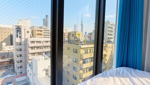 Hotellihuone, jossa on suuri ikkuna, sänky ja näkymä kaupungin siluettiin, johon kuuluu monenlaisia rakennuksia.