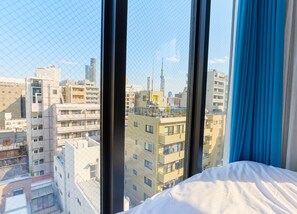 A hotel room with a large window, a bed, and a view of a cityscape with various buildings. - DeLCCS Ryogoku 2 (Sumida City)