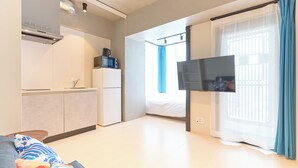 Moderni hotellihuone, jossa on pieni keittiö, taulutelevisio, sänky ja suuri ikkuna, jossa on verhot.