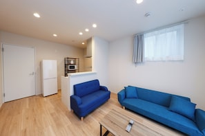 A compact living space with a kitchenette, two blue sofas, a white door, and a window with curtains. - La Terrasse Higashikomagata by Tranova 2 (Sumida City)