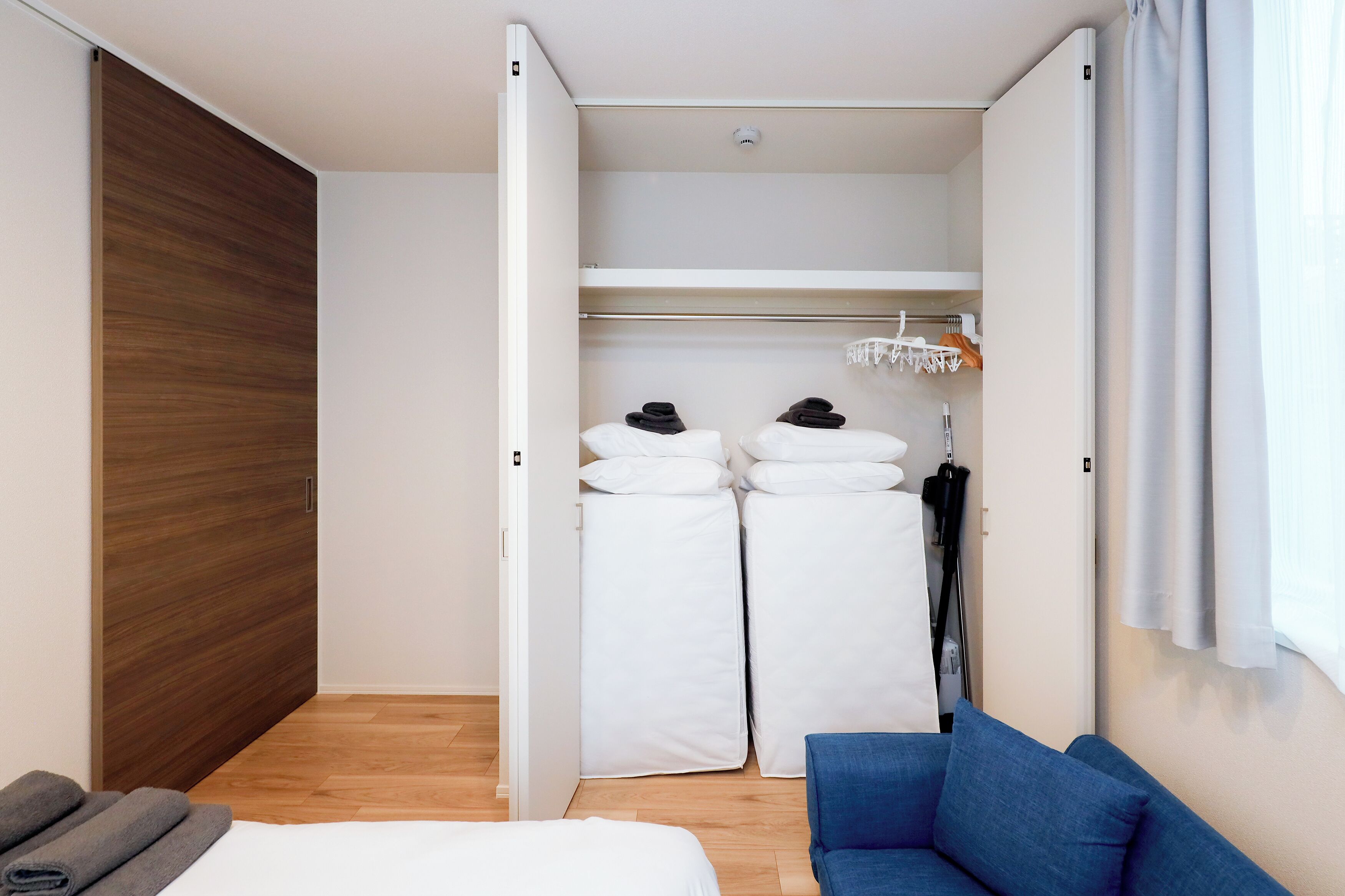 A compact room with a bed, a blue sofa, a wooden wardrobe, and a white closet with hanging clothes.