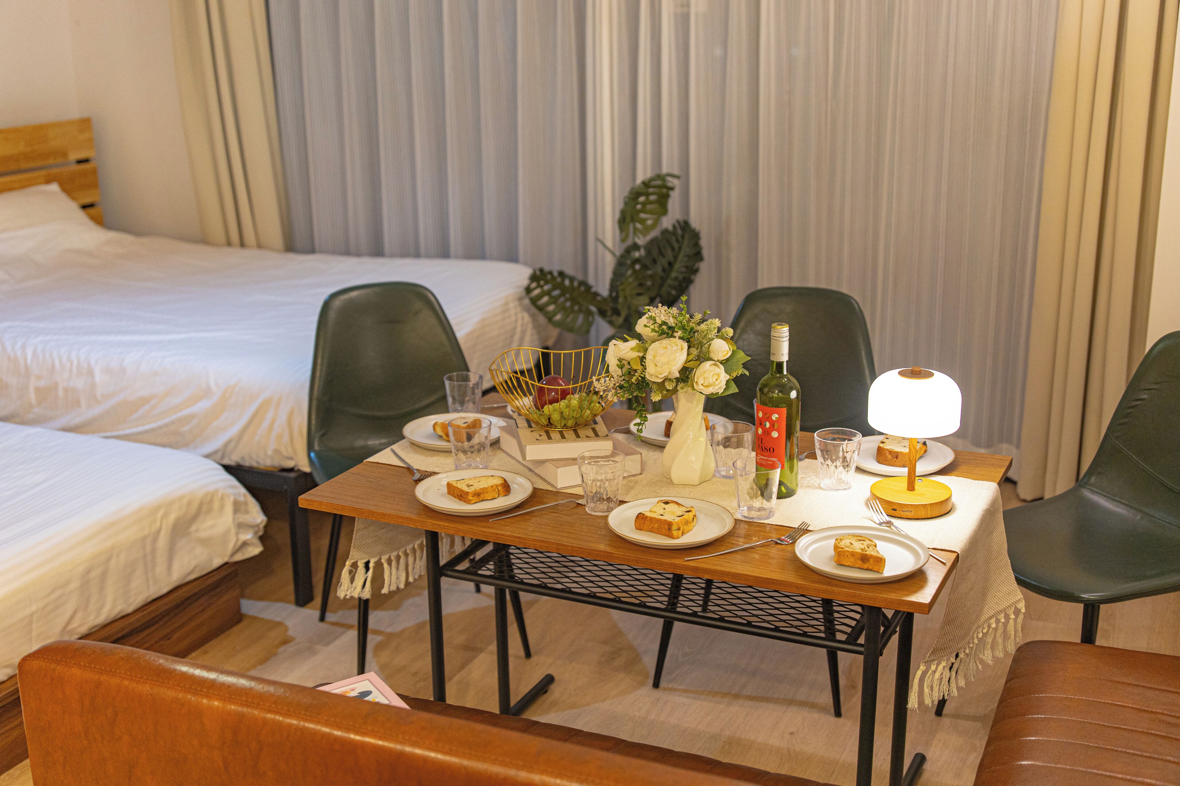 A hotel room with a neatly made bed, a dining table with plates of food, glasses, and a bottle of wine.