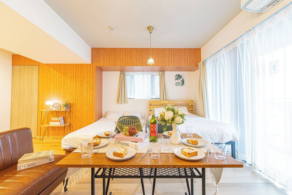 A hotel room with a bed, a dining table set with plates and glasses, a sofa, a small side table with a mirror and books, and a window with curtains.