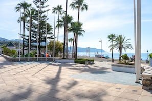 Ein gepflasterter Platz mit Palmen, Bänken und Blick auf Strand und Meer.