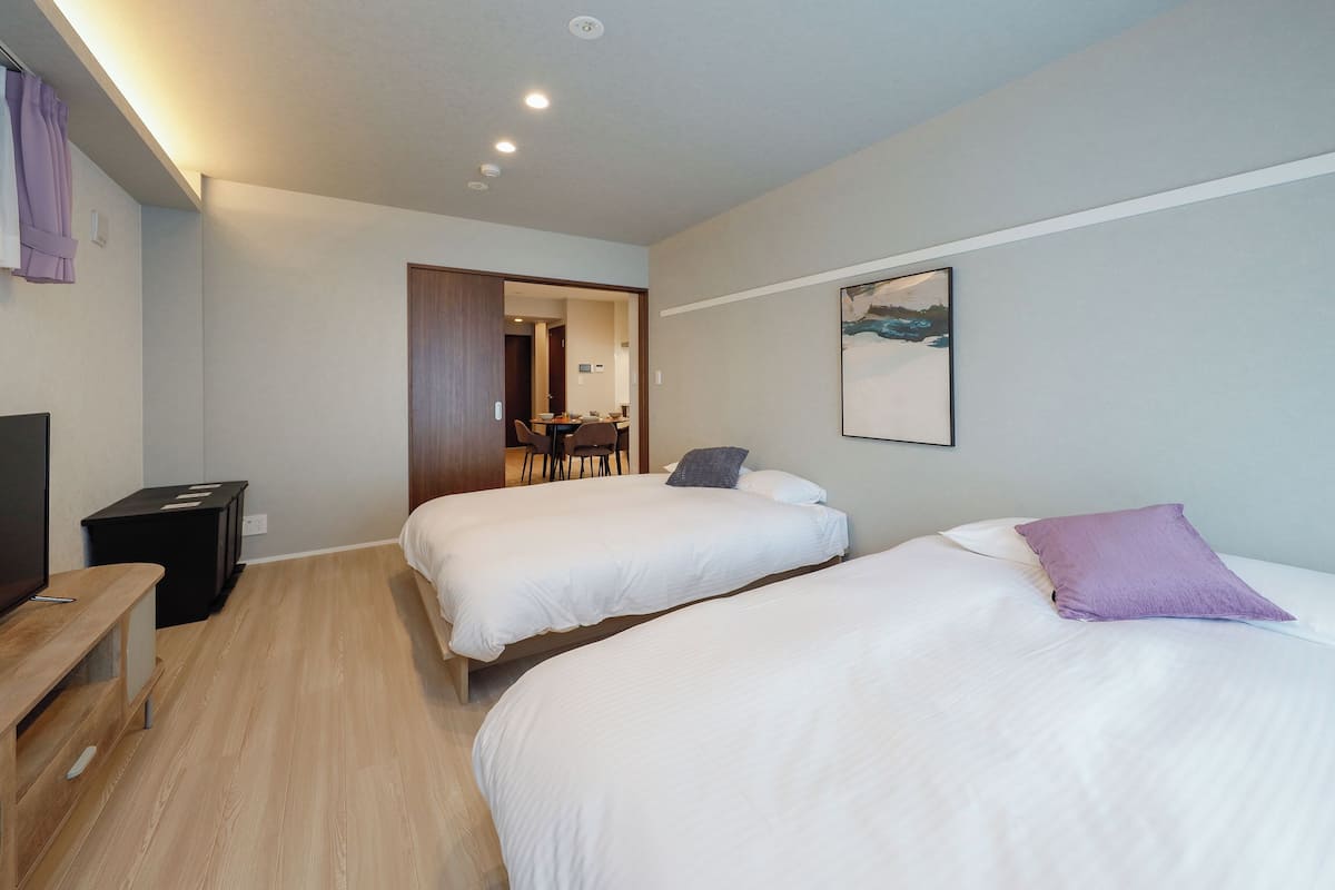 Quarto de hotel com duas camas, uma mesa de escritório, uma televisão e uma área de refeições visível através de uma porta aberta.