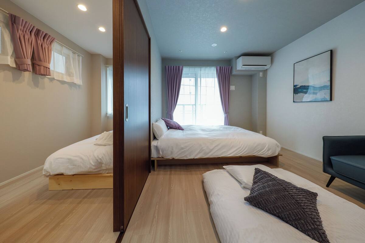 Quarto com duas camas, um sofá, uma janela com cortinas, ar condicionado e um quadro na parede.