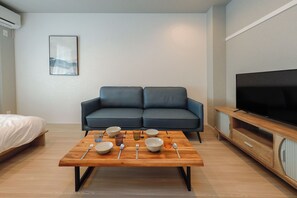 Un salon moderne avec un canapé, une table basse en bois sur laquelle sont posés des bols et des couverts, un téléviseur sur un meuble en bois, et un lit dans un coin.