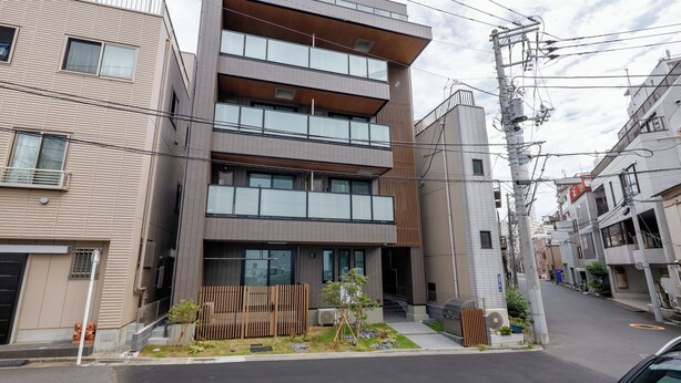 A modern multi-story building with a flat facade, large windows, and a small garden area with plants and a wooden fence. - AI Grandir Azumabashi 2 (Sumida City)