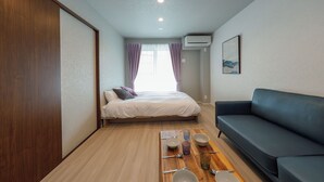 A room with a bed, a sofa, a dining table with cutlery, and an air conditioning unit. - AI Grandir Azumabashi 2 (Sumida City)