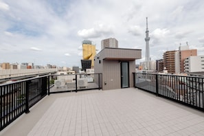 Terraço no telhado com piso de azulejos, guarda-corpo de metal preto e vista para o horizonte urbano, incluindo uma torre e edifícios.
