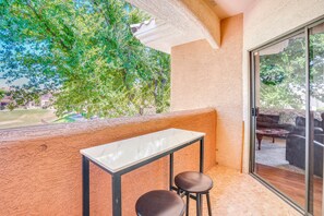 Ein Balkon mit Tisch und zwei Stühlen, eine Glastür zu einer Außenbereich-Sitzgelegenheit und Blick auf Bäume und Gebäude.