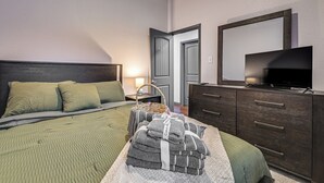 Ein ordentlich bezogenes Bett mit grüner Bettwäsche, eine dunkle Holzkommode mit einem Flachbildfernseher und ein Korb gefalteter Handtücher.