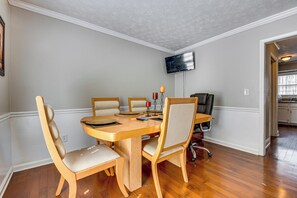 Un coin repas avec une table en bois, quatre chaises, un téléviseur fixé au mur et une cuisine visible au fond.