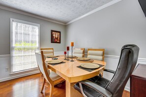 Uma sala de jantar com uma mesa de madeira posta para quatro pessoas, quatro cadeiras, uma janela com persianas e um quadro na parede.