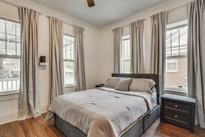 Un dormitorio con cama, mesita de noche y ventanas amplias con cortinas.