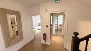 Un pasillo con suelo de madera, un espejo, un extintor y una puerta que da a otra habitación.