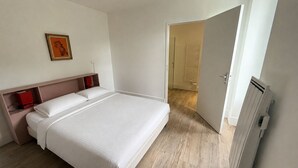 Un dormitorio con cama, mesita de noche, lámpara roja, un cuadro enmarcado y una puerta que da a otra habitación.
