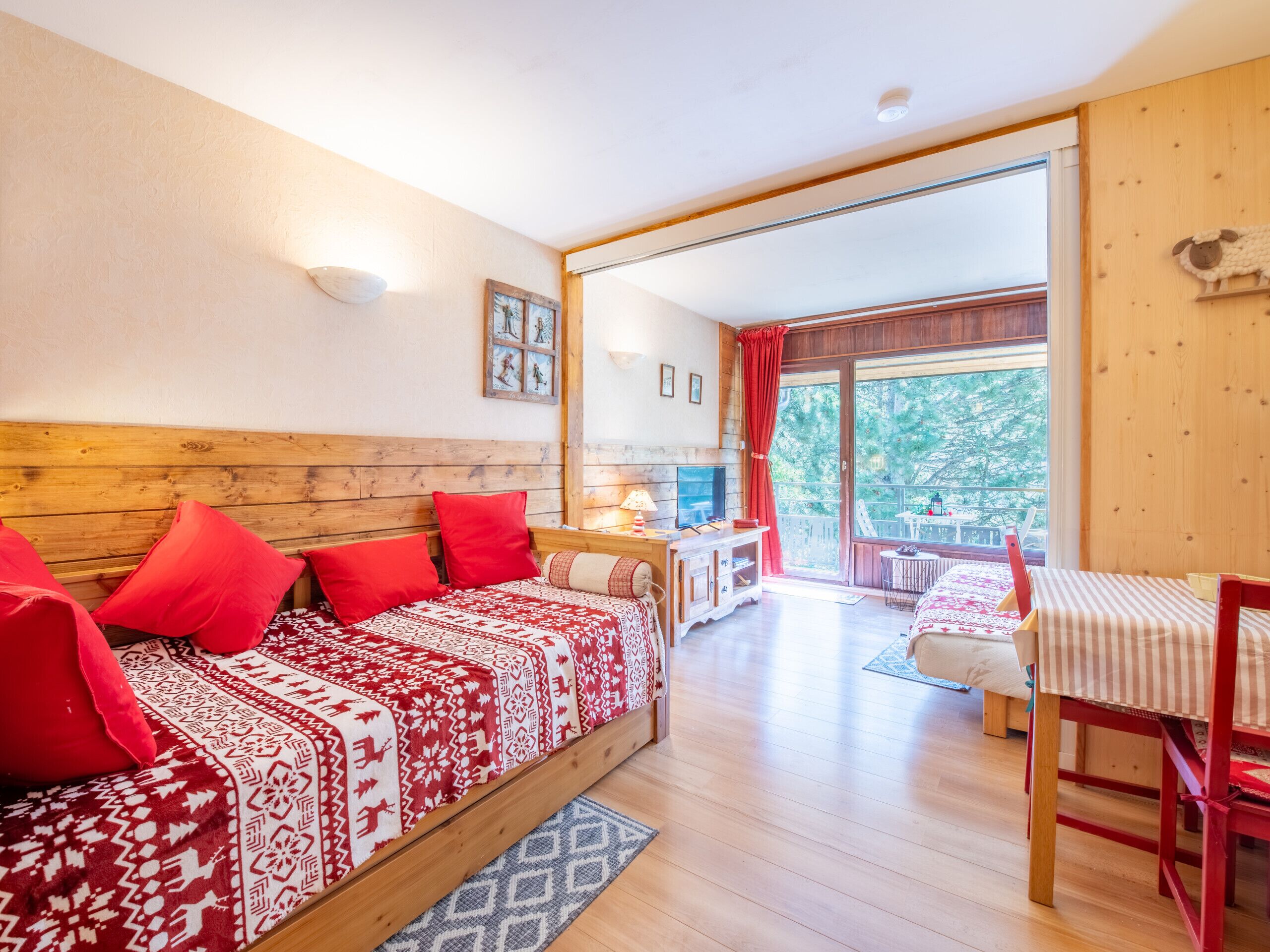 Een gezellige kamer met een houten bed, een klein tafeltje met stoel, en een groot raam met uitzicht op bomen.