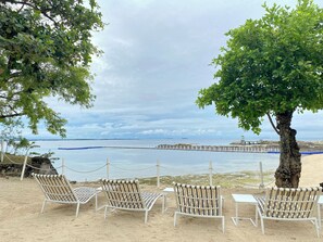 Ein Strand mit weißen Liegestühlen und einem Baum, mit Blick auf ein ruhiges Meer und einen Steg.