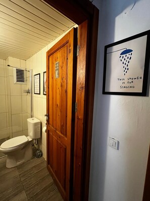 Une salle de bain avec une porte en bois, des toilettes, une douche et un panneau encadré accroché au mur.