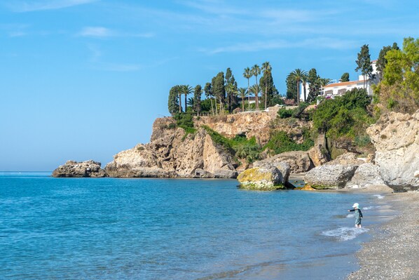 A coastal landscape with rocky terrain, a clear blue sky, and a building on a hill. - Tahiti 2 7 by Casasol (Nerja)