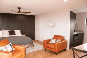 A modern bedroom with a bed, two chairs, a desk, a ceiling fan, and a kitchenette.