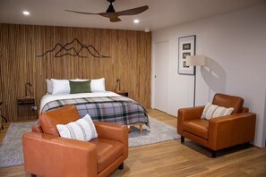 A modern bedroom with a bed, two leather armchairs, a wooden slatted wall, a ceiling fan, and a bedside table.