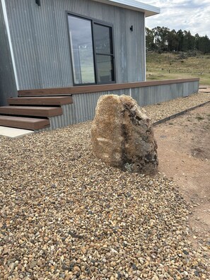 A modern building with a metal exterior, a large rock in the foreground, and a gravel area.