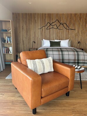A modern bedroom with a leather armchair, a bed with a plaid blanket, a wooden slat wall, and a bookshelf.
