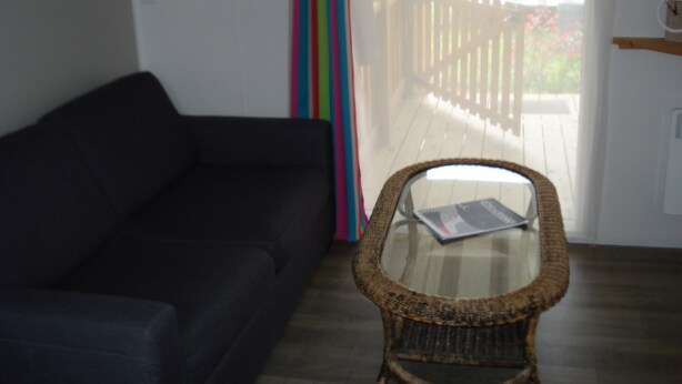 Una habitación con un sofá de color oscuro, una silla de mimbre con superficie de vidrio, una terraza de madera visible a través de una ventana y una pared blanca con un reloj.