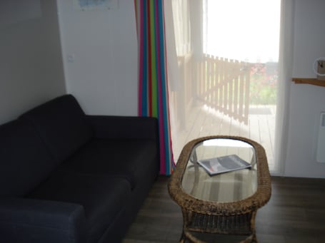 A room with a dark-colored sofa, a wicker chair with a glass top, a wooden deck visible through a window, and a white wall with a clock.. 2-bedroom mobile home in awesome Saint-Hilaire-de-Riez with AC