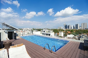 Una piscina en la azotea con vistas al horizonte de la ciudad, una terraza de madera y una zona junto a la piscina con sillones de descanso.