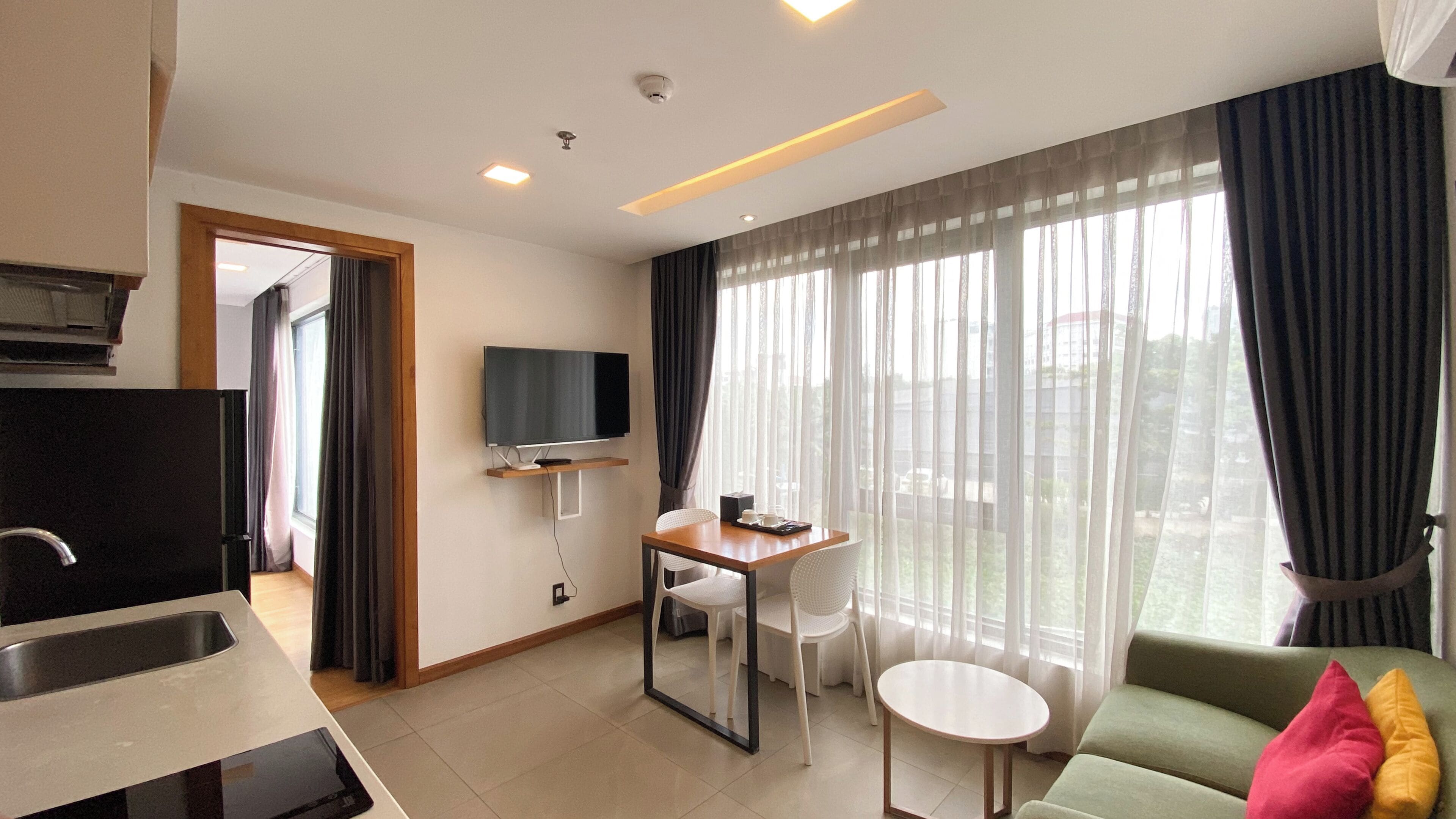 Una habitación de hotel moderna con kitchenette, un televisor de pantalla plana montado en la pared, un área de comedor con mesa y sillas, y un sofá con cojines.