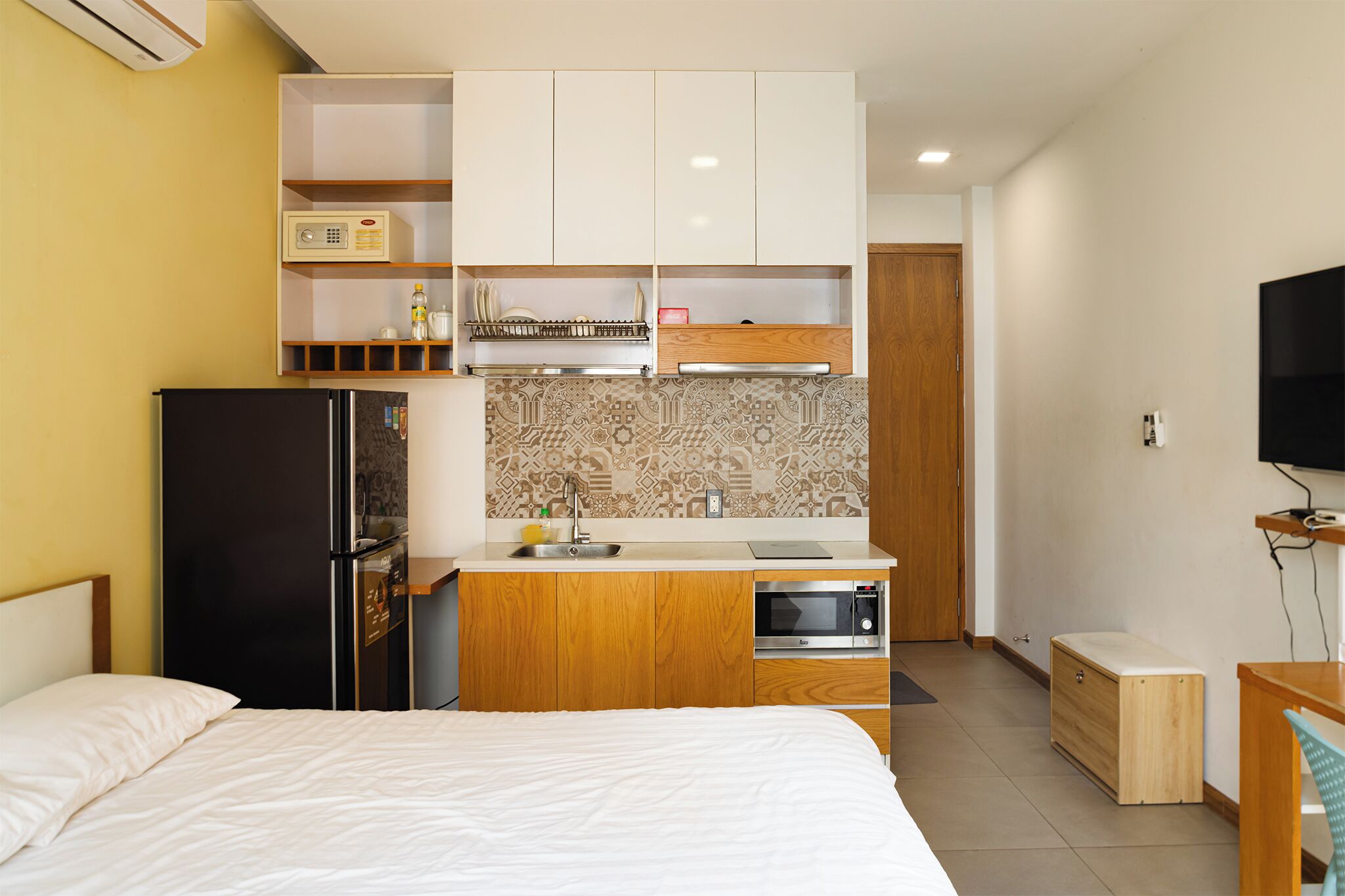 A compact hotel room with a bed, a kitchenette with a refrigerator, microwave, and sink, and a television on the wall.