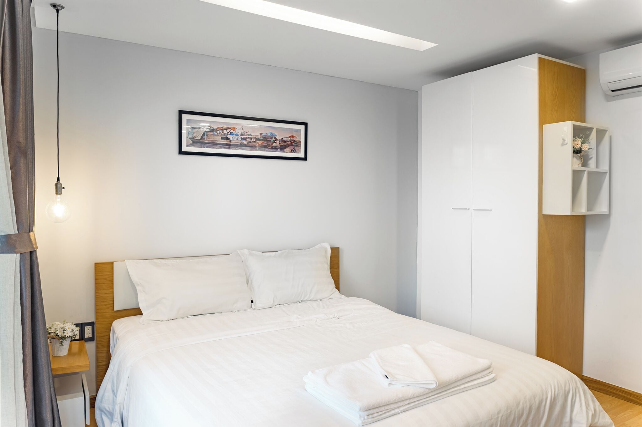 Quarto de hotel com uma cama, mesa de cabeceira, quadro na parede, guarda-roupa e uma pequena estante com flores.
