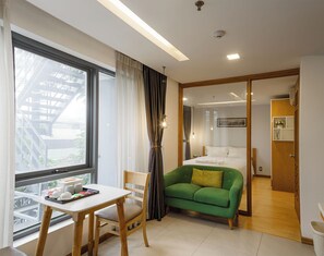 Quarto de hotel com cama, sofá verde, mesa de jantar com cadeiras, janela com cortinas e vista para um edifício.