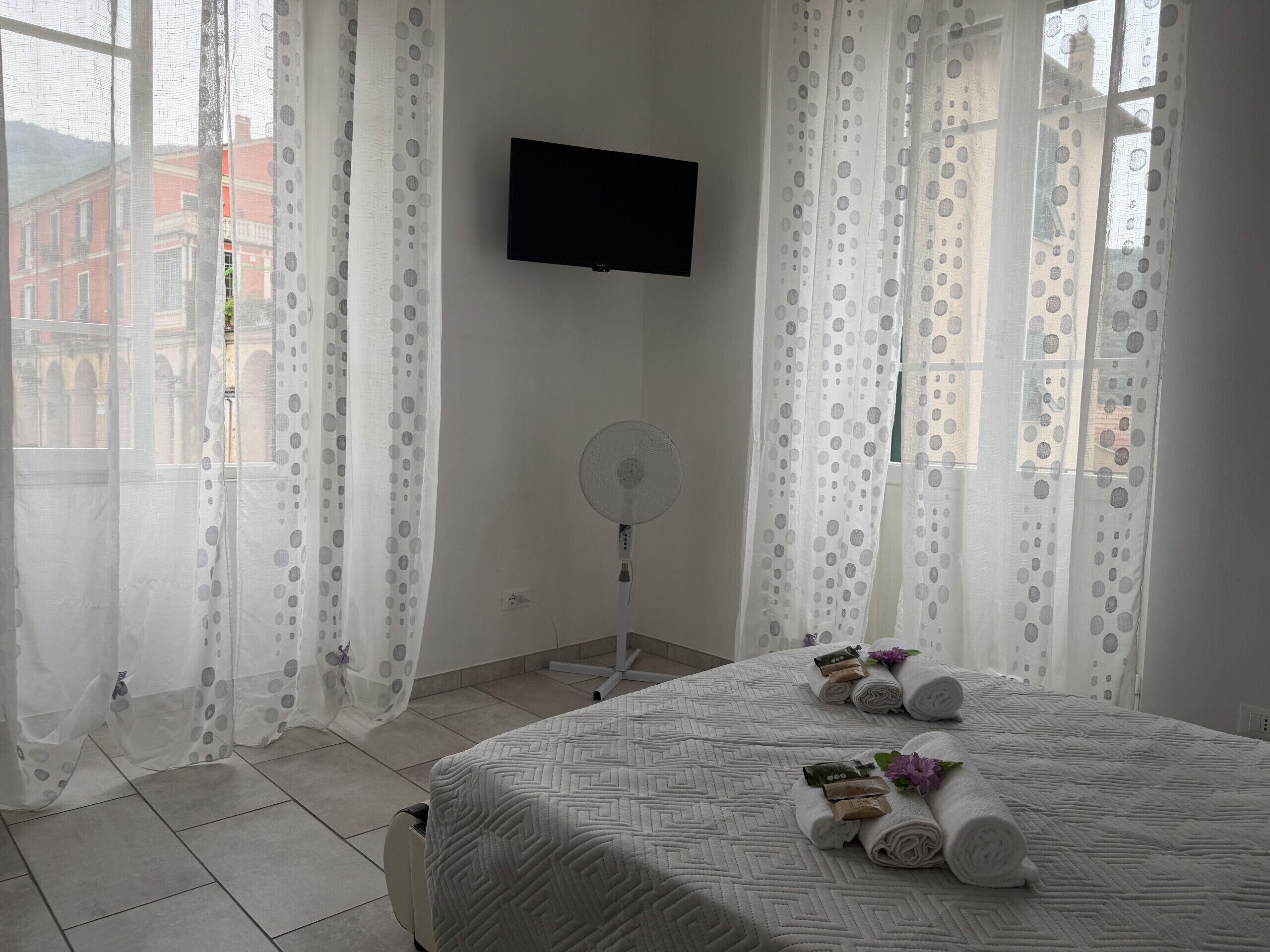 Ein Hotelzimmer mit einem Bett, einem Flachbildfernseher, einem Standventilator und einem Fenster mit durchsichtigen Vorhängen.