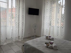 A hotel room with a bed, a flat-screen TV, a standing fan, and a window with sheer curtains. - Casa Nina - In downtown Pontedassio, 5 minutes from Imperia, by HOST4U (Pontedassio)